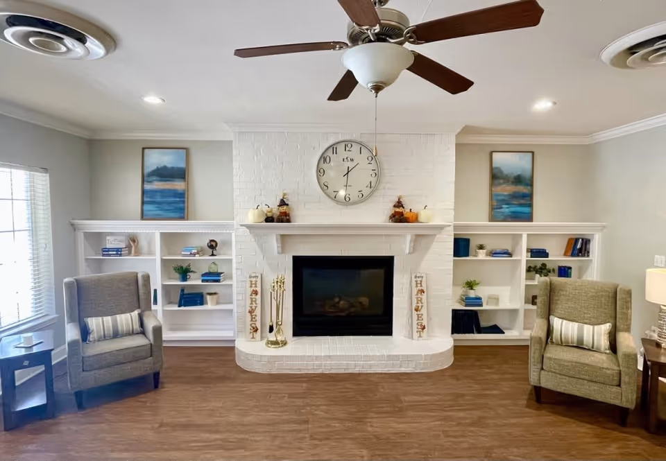 A cozy living room with a white brick fireplace in the center, flanked by built-in white bookshelves filled with books and decorative items. Two comfortable armchairs with striped pillows are positioned on either side of the fireplace. Above the fireplace is a round wall clock, and two framed landscape paintings hang on the walls above the bookshelves. A ceiling fan with wooden blades is mounted on the ceiling, and the room has wood flooring and soft lighting.