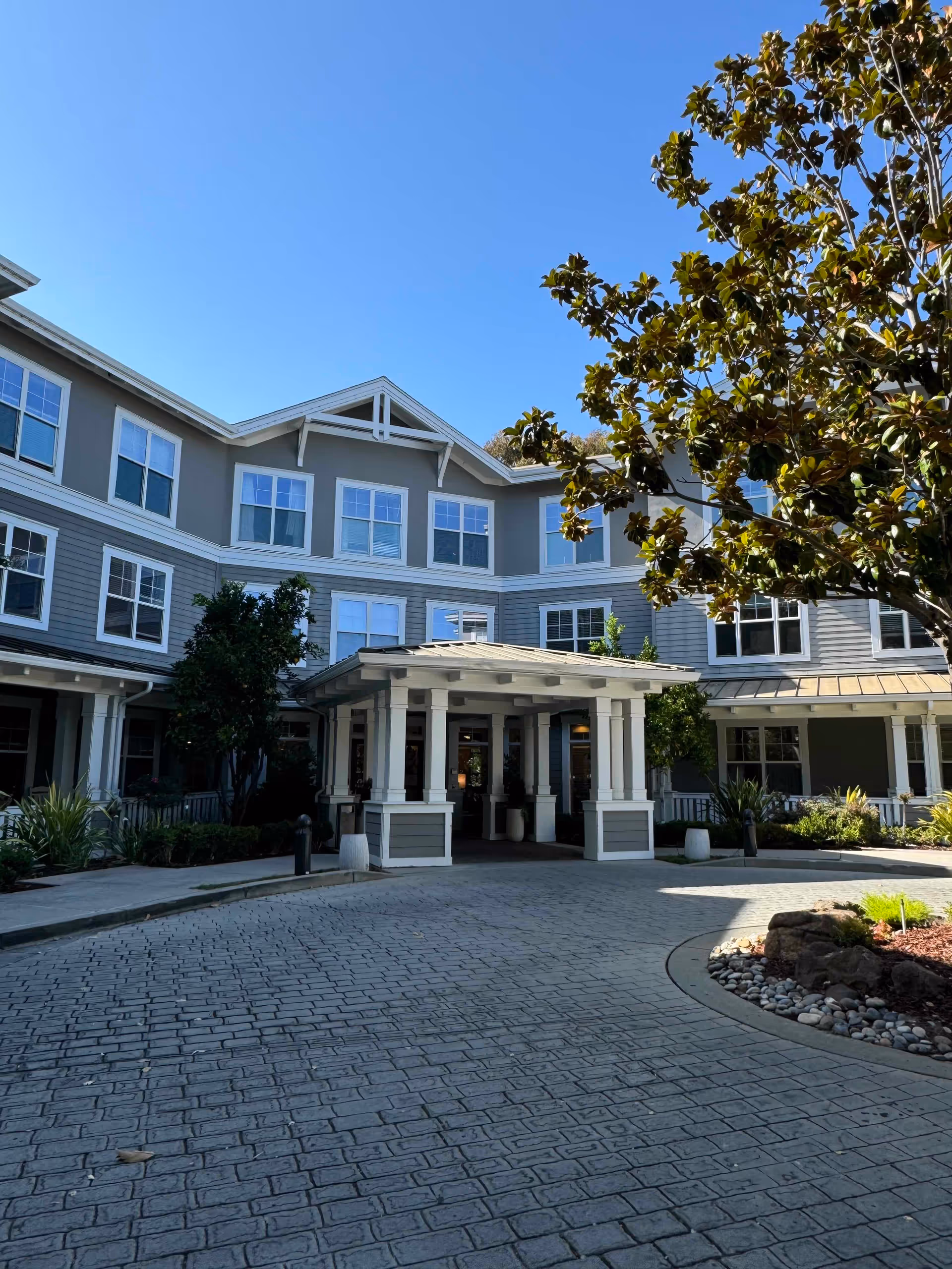 Front entrance of a multi-story senior living building with a covered porte-cochere, windows, landscaping and a tree.