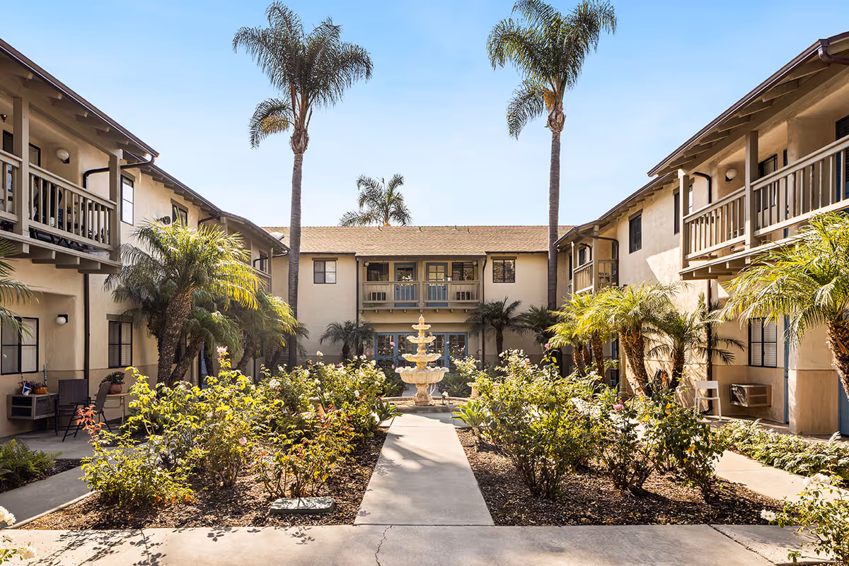 Outdoor courtyard area of a senior living facility with a central walkway leading to a multi-tiered fountain surrounded by flower beds and palm trees. The courtyard is flanked by two-story beige buildings with balconies and windows under a clear blue sky.
