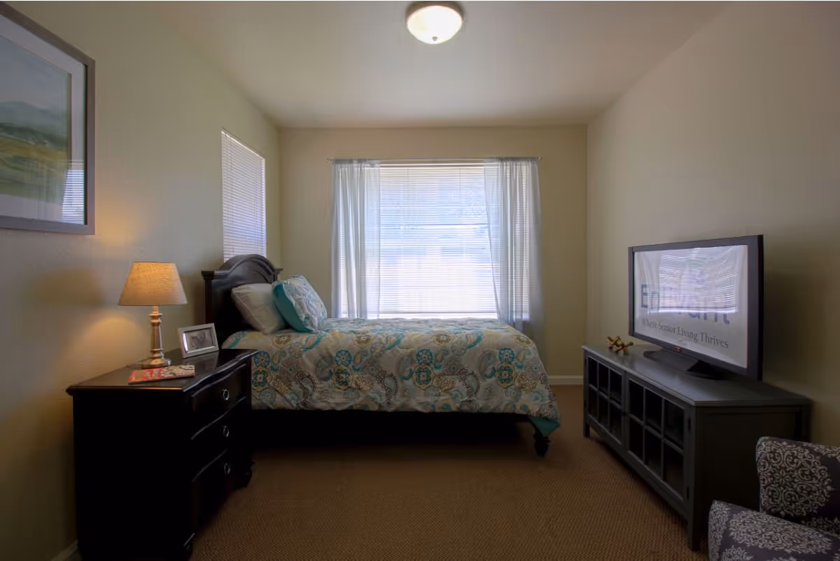 Well-lit furnished bedroom with a bed centered under a window, a nightstand and lamp to the left, and a TV on a console to the right.