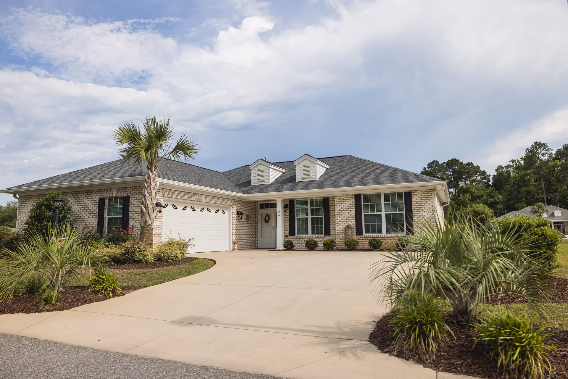 Single-story brick house with a gray shingled roof, white garage door, and a concrete driveway. The front yard features landscaped plants including palm trees and bushes under a partly cloudy sky.
