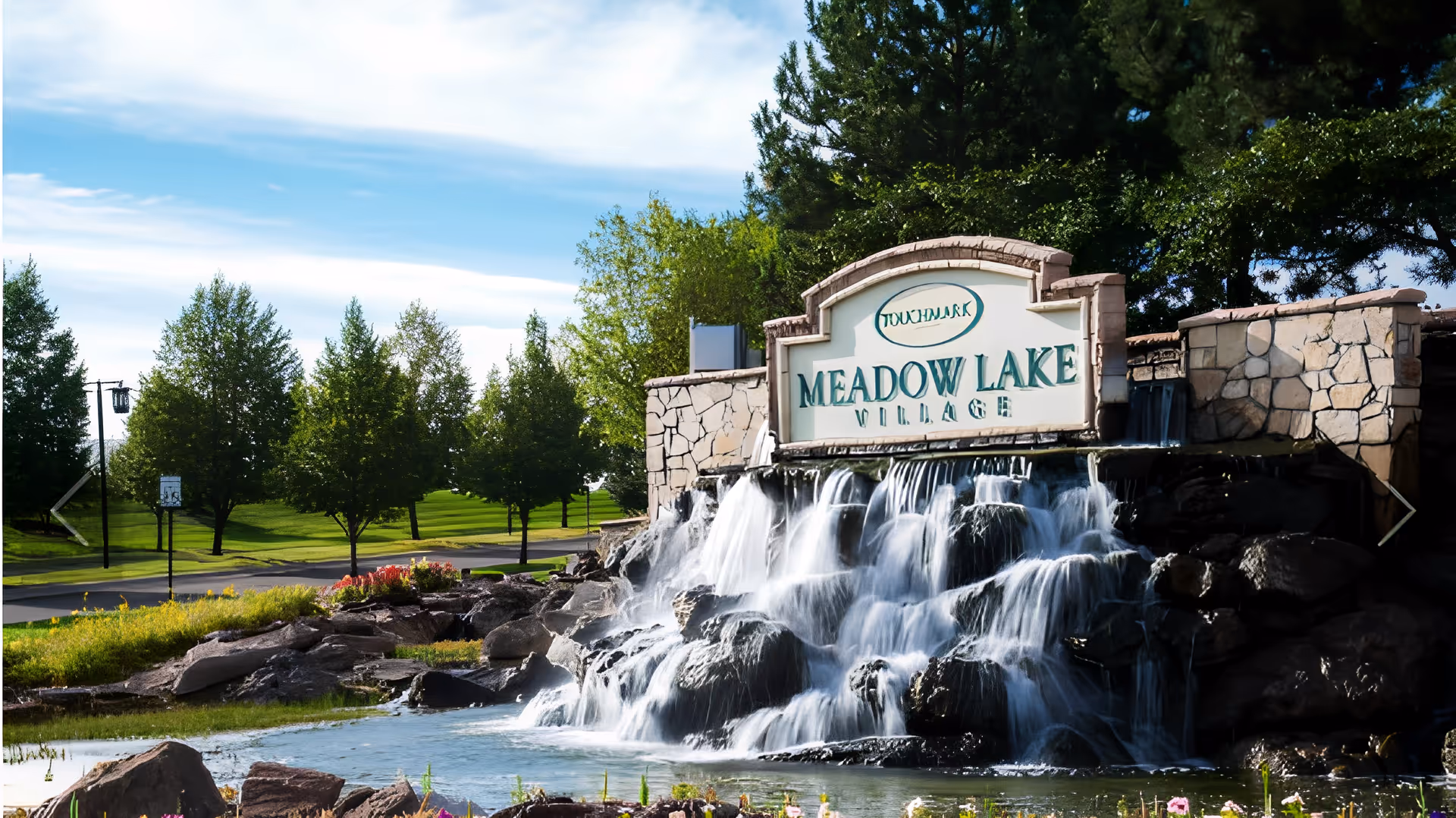 Stone entrance sign reading 'Meadow Lake Village' with a cascading waterfall and landscaped grounds.