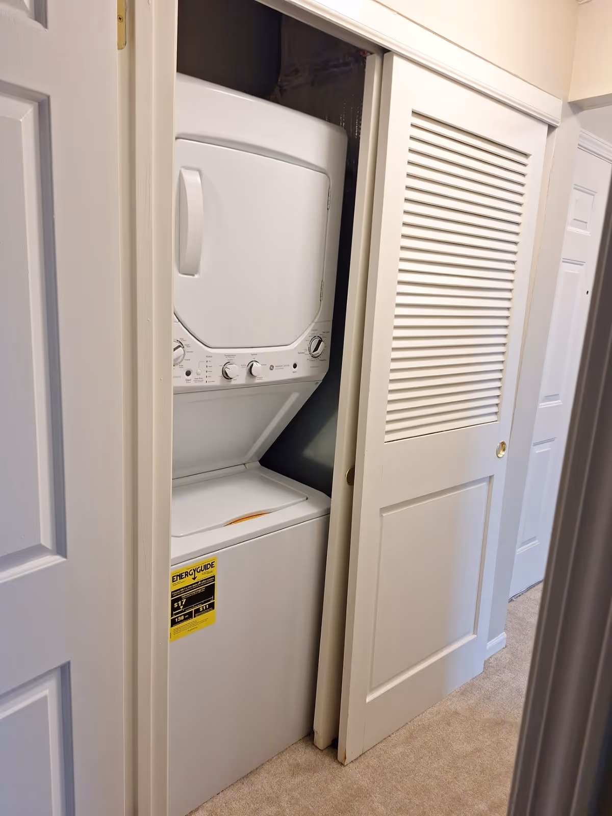 A stacked white washer and dryer unit inside a small closet with a sliding louvered door partially open, located in a hallway with beige carpet and white walls.
