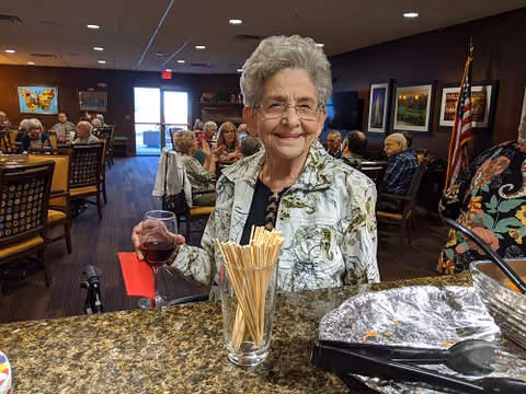 An elderly woman with short gray hair and glasses is smiling and holding a glass of red wine while standing at a counter in a dining area. Behind her, several other elderly people are seated at tables, engaged in conversation. The room has framed artwork on the walls and an American flag in the background.