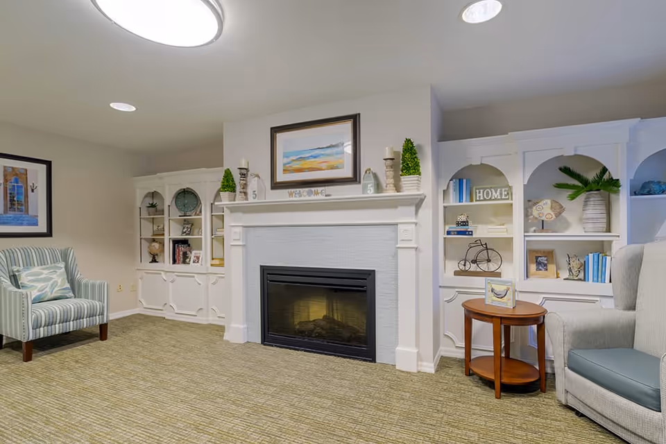 A cozy living room area with a fireplace in the center, flanked by white built-in shelves decorated with books, plants, and ornaments. There is a striped armchair with a patterned cushion on the left and a gray armchair with a blue cushion on the right next to a small wooden round table. The walls are light-colored, and the floor is carpeted.