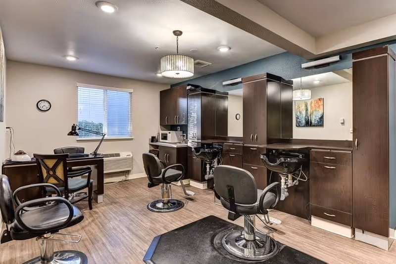 Interior salon area with styling chairs, shampoo sinks, mirrors, and dark cabinetry in a senior living facility.