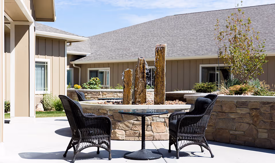 Outdoor patio area at a senior living facility with a round glass-top table and two black wicker chairs. In the background, there is a stone water fountain with three vertical stone pillars with water flowing down. The building exterior is beige with white-trimmed windows and a gray shingled roof. There are some plants and small trees around the patio area.