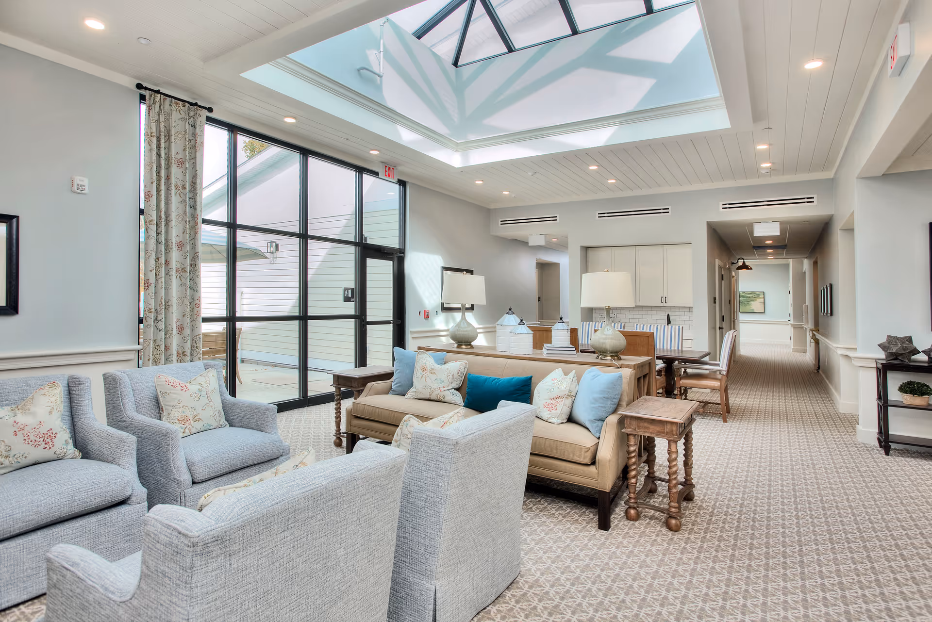 A bright and spacious senior living common area with comfortable seating including light blue armchairs and a beige sofa adorned with decorative pillows. The room features a large skylight ceiling, patterned carpet, and large glass doors leading to an outdoor patio. There are two table lamps on a console table behind the sofa and a dining table with chairs in the background.