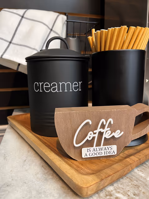 A close-up of a wooden tray holding a black container labeled 'creamer', a black cup filled with wooden stir sticks, and a wooden coffee cup-shaped sign that reads 'Coffee is always a good idea'. A white and gray checkered cloth is hanging in the background.