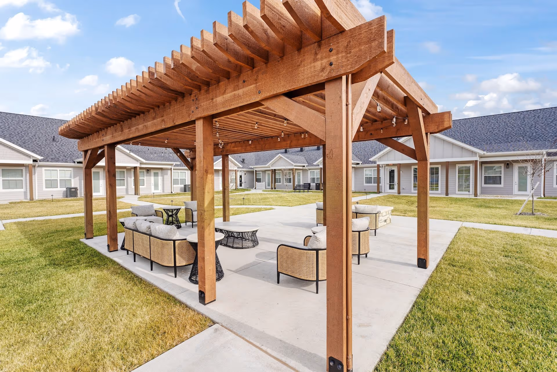 Outdoor seating area under a wooden pergola with cushioned chairs and tables, surrounded by a grassy courtyard and single-story residential buildings under a partly cloudy sky.