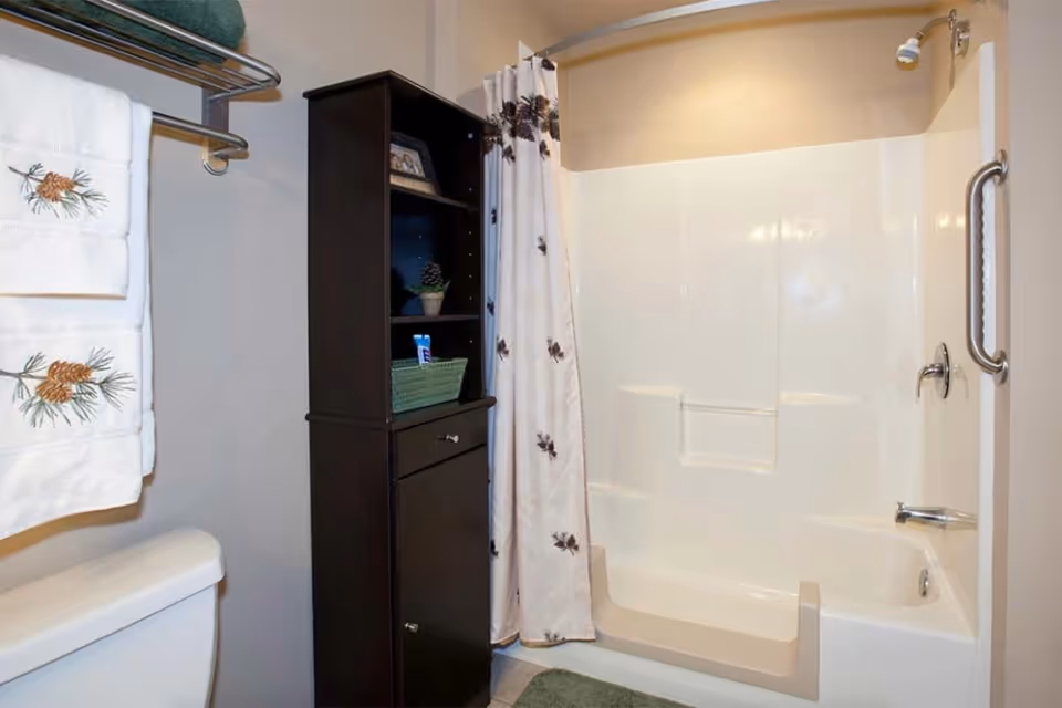 A bathroom with a white bathtub and shower combination featuring a built-in seat and grab bar. There is a shower curtain with a pinecone pattern, a dark wooden storage cabinet with shelves and drawers, a towel rack holding white towels embroidered with pinecones, and a toilet partially visible in the foreground.