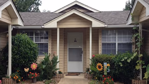 Front exterior view of a single-story residential building with beige siding, a white door, and large windows on either side. The entrance is framed by a small porch with a peaked roof, surrounded by green shrubs and colorful flowers.