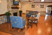 A cozy living room area in a nursing and rehabilitation facility featuring wooden flooring, a stone fireplace with a TV mounted above it, two blue patterned armchairs, a beige sofa, a wooden coffee table with decorative items, and wall decorations. The room is well-lit with recessed lighting and has an open layout with a hallway visible in the background.