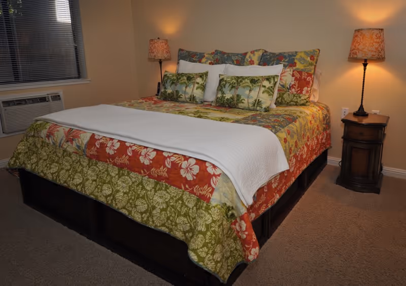 A cozy bedroom with a bed featuring a colorful floral and palm tree patterned bedspread, multiple pillows, two bedside tables each with a lamp, a window with blinds, and a wall-mounted air conditioning unit.