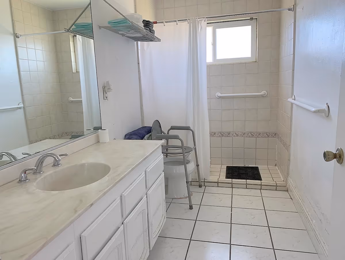 A clean bathroom with white tiled floor and walls. There is a sink with a countertop and a large mirror above it on the left side. A commode with a gray walker frame around it is positioned near a walk-in shower with a white curtain and a small window above. Towels are neatly stacked on a metal rack above the commode. Grab bars are installed on the walls near the shower and door for safety.