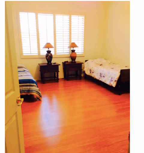 Bright bedroom with two twin beds, two bedside tables topped with lamps, a large shuttered window, and wood flooring.