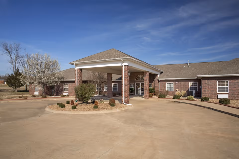 Front exterior view of Oak Manor Nursing Center, a single-story brick building with a covered entrance supported by brick columns, surrounded by a circular driveway and landscaped with small bushes and trees under a clear blue sky.