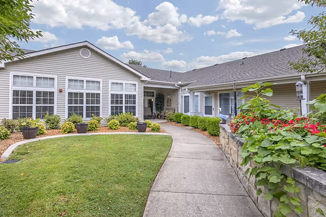 Exterior view of a single-story senior living facility building with beige siding, multiple large windows, and a gray shingled roof. A concrete walkway curves through a well-maintained lawn and landscaped garden beds with shrubs and flowers. The sky is partly cloudy with blue patches visible.