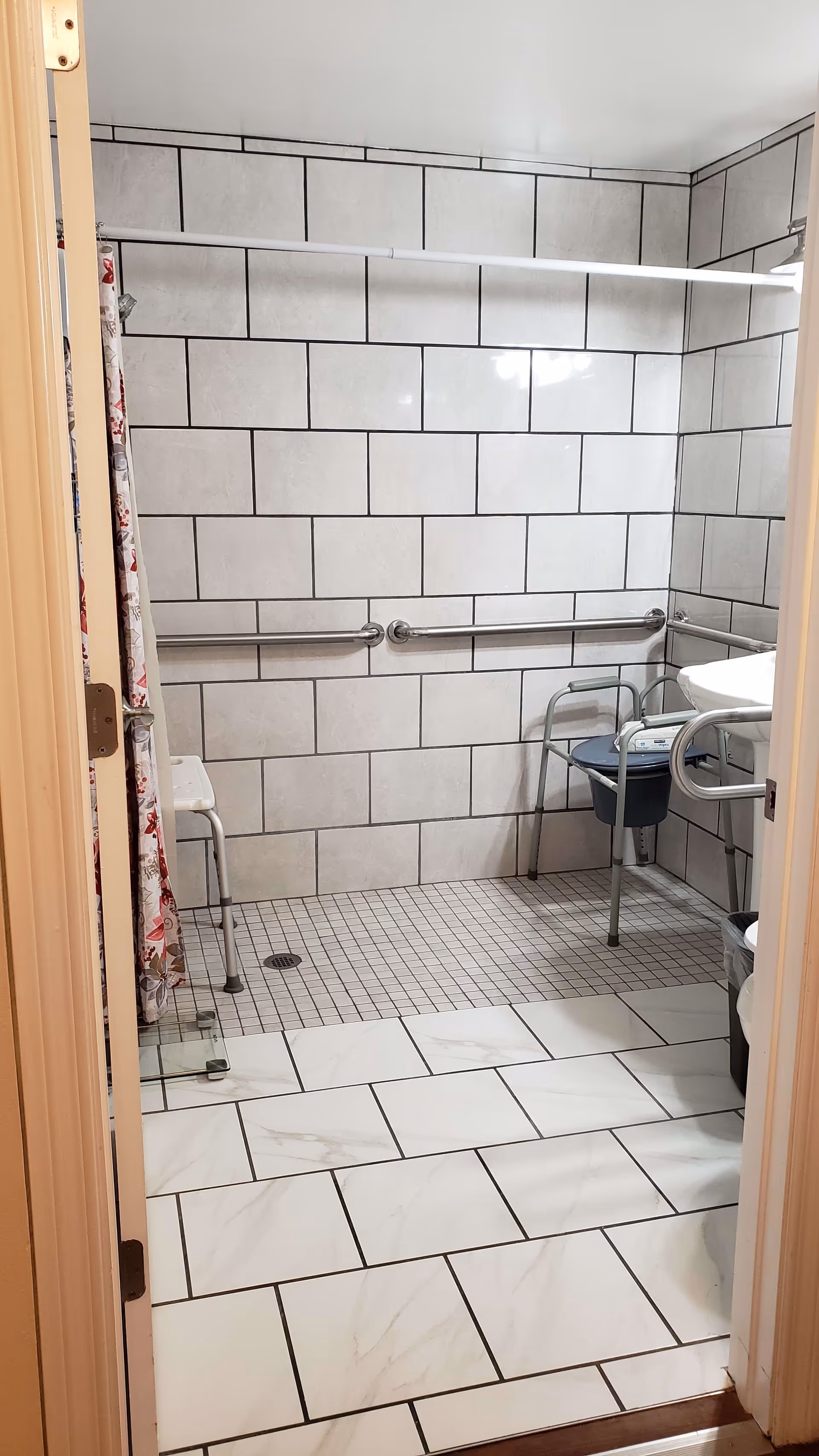 View into a tiled bathroom with a walk-in shower area featuring a shower chair, grab bars on the walls, and a shower curtain. The floor has large white tiles with gray veining, and the shower area has smaller square tiles. There is a sink with a grab bar and a small waste bin visible.