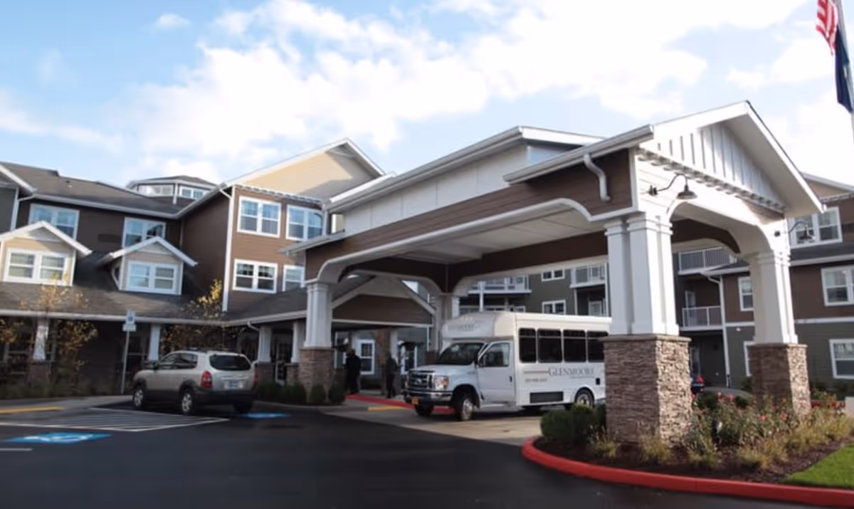 The front entrance and covered porte-cochère of Glenmoore Gracious Retirement Living with a shuttle van, parked cars, and the multi-story building facade.
