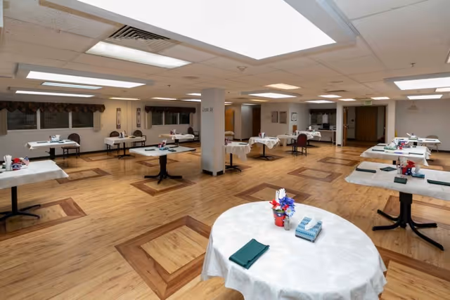 A spacious dining room with multiple round and rectangular tables covered with white tablecloths. Each table is set with green napkins, tissue boxes, and small flower arrangements. The room has wooden flooring with decorative square patterns and is illuminated by ceiling lights. There are windows with valances along the walls and a few chairs placed around the tables.