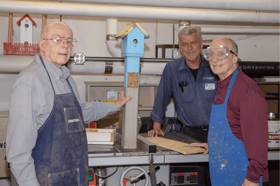 Three elderly men in a workshop setting. One man on the left is wearing glasses and a blue apron labeled 'CRAFTSMAN,' holding a tall birdhouse painted blue with a yellow roof. The man in the middle is standing behind a workbench, wearing a blue striped shirt with a name tag. The man on the right is wearing safety goggles, a maroon shirt, and a blue apron, smiling at the camera. Various tools and workshop equipment are visible in the background.