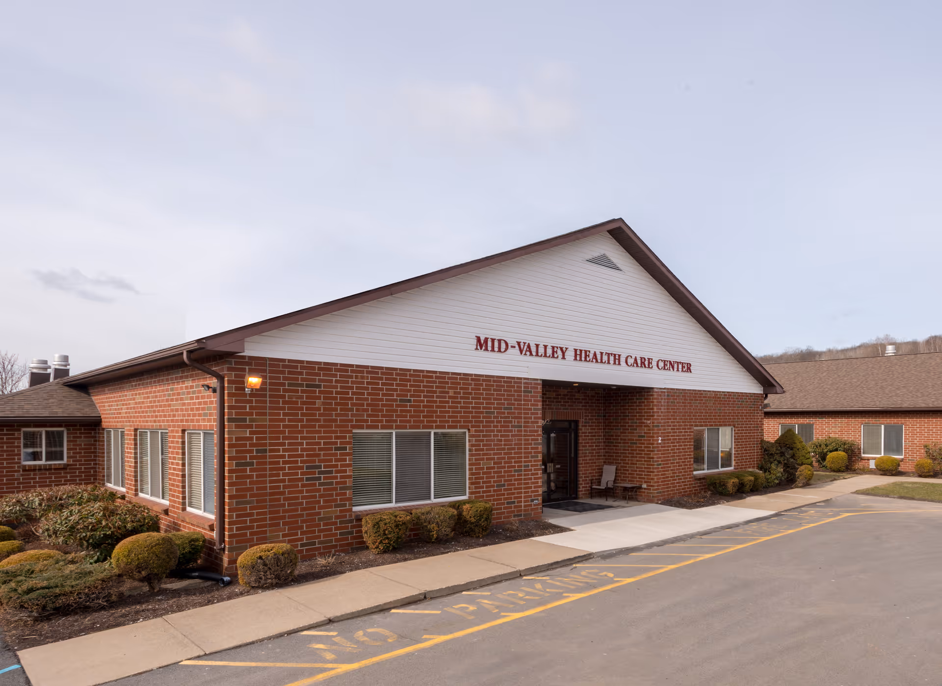 Exterior view of Mid-Valley Health Care Center, a single-story brick building with a white triangular gable above the entrance. The building is surrounded by small bushes and a paved driveway with a 'No Parking' zone marked in yellow.