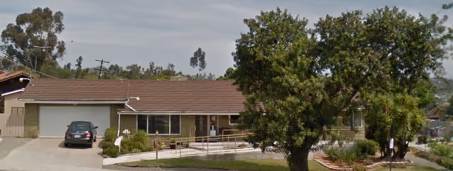 Single-story residential building with a brown tiled roof, a garage on the left with a parked car, a ramp leading to the front entrance, and trees and shrubs in the front yard under a partly cloudy sky.