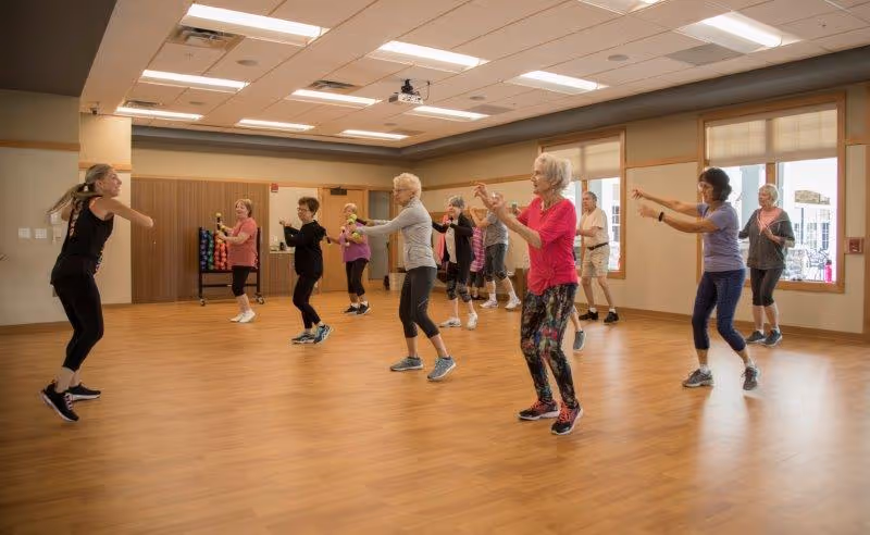 A group of elderly women participating in a fitness or dance class in a spacious room with wooden floors and large windows. An instructor leads the class from the front left side.