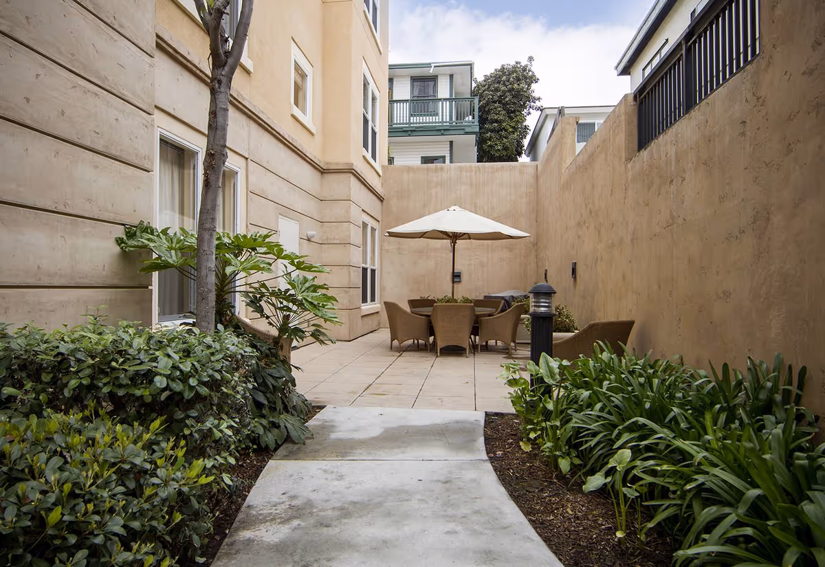 Enclosed courtyard with a tiled patio, round table and wicker chairs under a beige umbrella, flanked by plants and tall stucco walls.