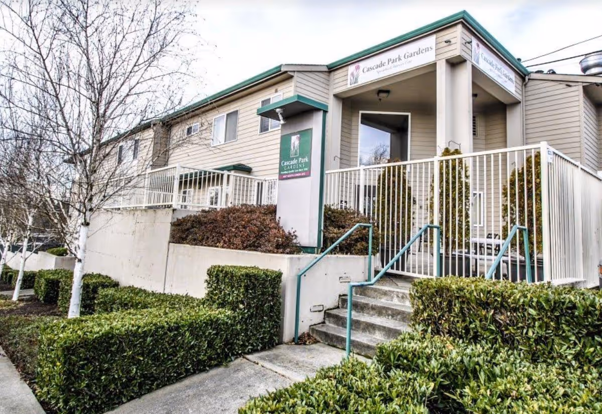Exterior view of Cascade Park Gardens senior living facility showing the building entrance with stairs, railings, bushes, and leafless trees in the foreground.