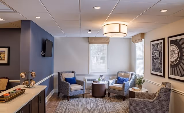 Bright seating area with upholstered armchairs around a small round table, framed wall art, and a mounted TV in a senior living facility.