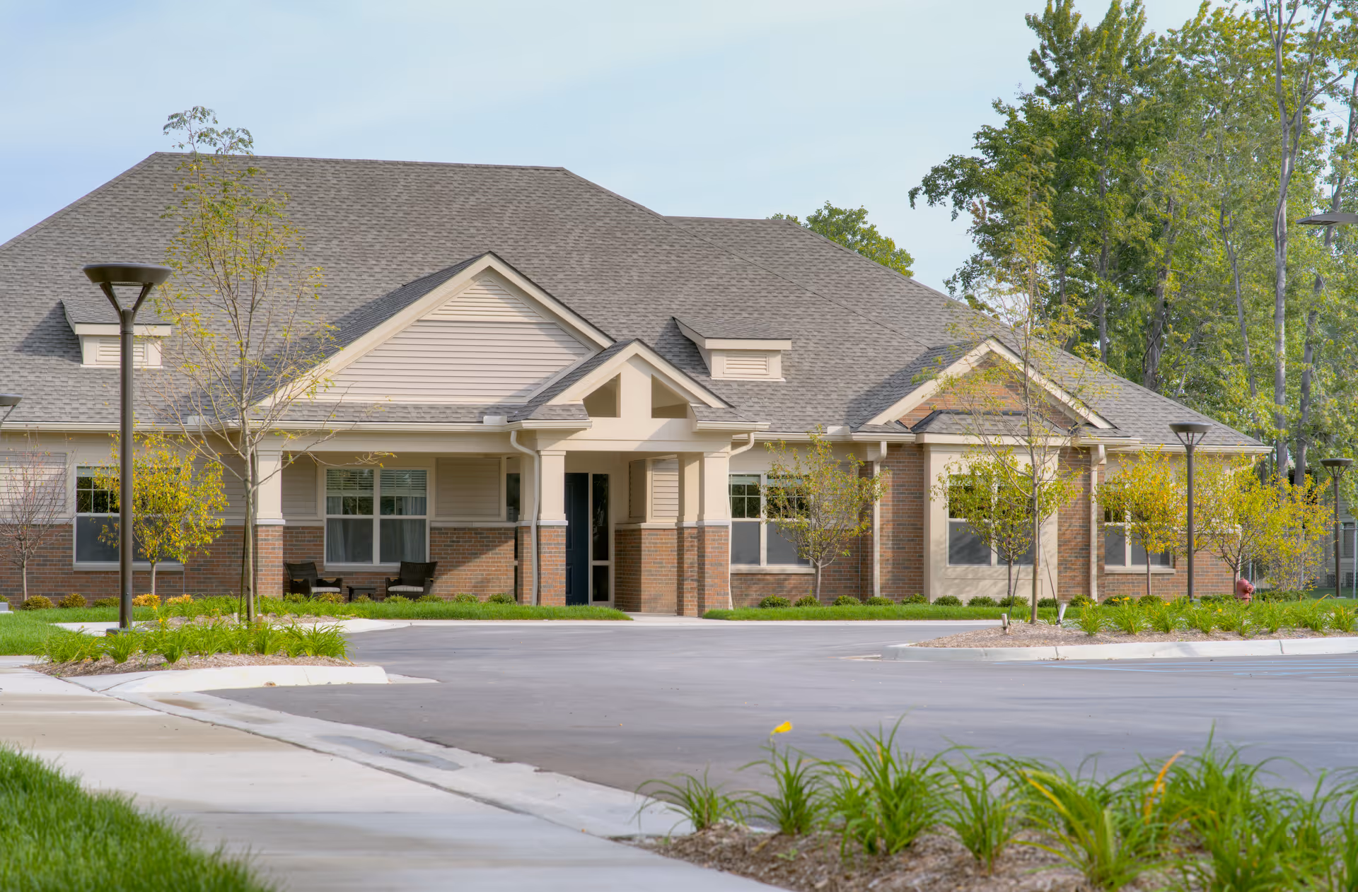 Exterior view of a single-story senior living facility building with a pitched roof, brick and siding facade, surrounded by young trees and landscaping, with a paved driveway and sidewalk in front.