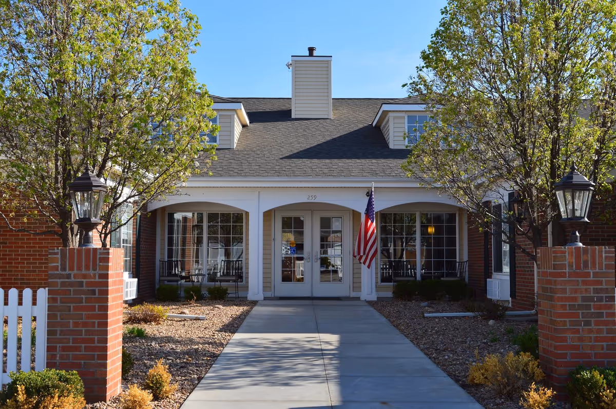 Front exterior view of a single-story building with a central entrance featuring double glass doors, flanked by large windows. The building has a gray shingled roof with dormer windows and a chimney. There are two brick pillars with lanterns on either side of the concrete walkway leading to the entrance. Trees with green leaves and landscaping with rocks and shrubs surround the walkway. An American flag is displayed near the entrance.