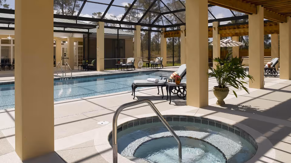 Indoor pool area with a hot tub in the foreground, lounge chairs with towels and a small table with flowers, potted plants, and a screened enclosure allowing natural light to enter.