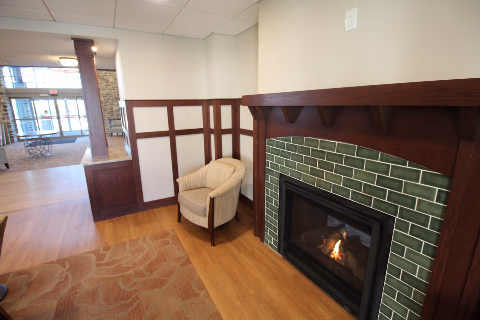 A cozy corner in a senior living facility featuring a beige upholstered armchair next to a fireplace with green tile surround and dark wood mantel. The room has wood flooring, a patterned area rug, and wood paneling on the walls. In the background, there is a view of a spacious lobby area with large windows and seating.