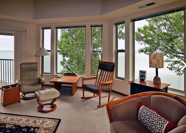 A cozy living room with large windows showing a view of trees and water outside. The room features a beige leather armchair with matching ottoman, a wooden rocking chair with blue cushions, a wooden side table with a lamp, and a cushioned armchair with a decorative pillow. The floor is carpeted and there is a patterned rug partially visible.