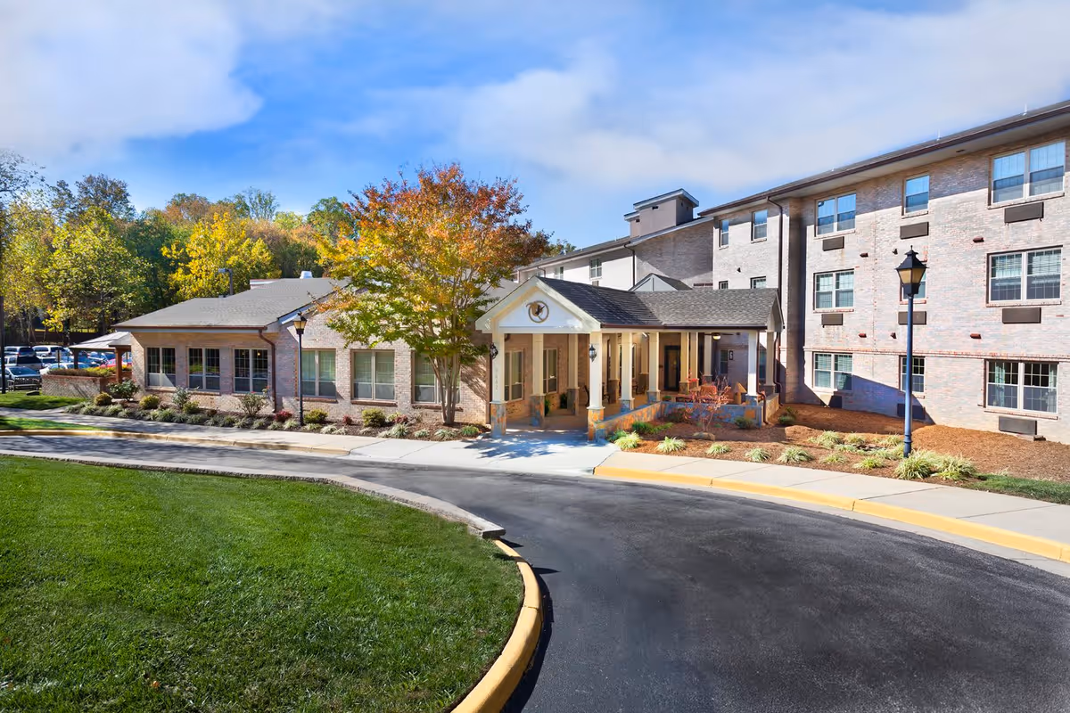 Exterior view of Heatherwood Retirement Community showing a multi-story brick building with large windows, a covered entrance with columns, landscaped grounds with green grass, trees with autumn foliage, and a curved driveway.