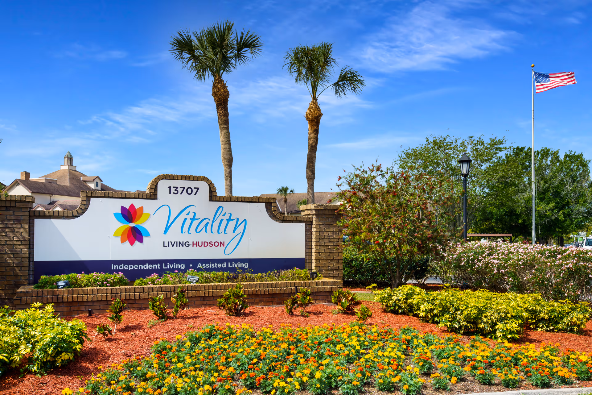 Outdoor view of the entrance sign for Vitality Living Hudson, a senior living facility. The sign is surrounded by landscaped flower beds with colorful flowers and shrubs, two tall palm trees behind the sign, and an American flag flying on a flagpole to the right. The sky is clear and blue.