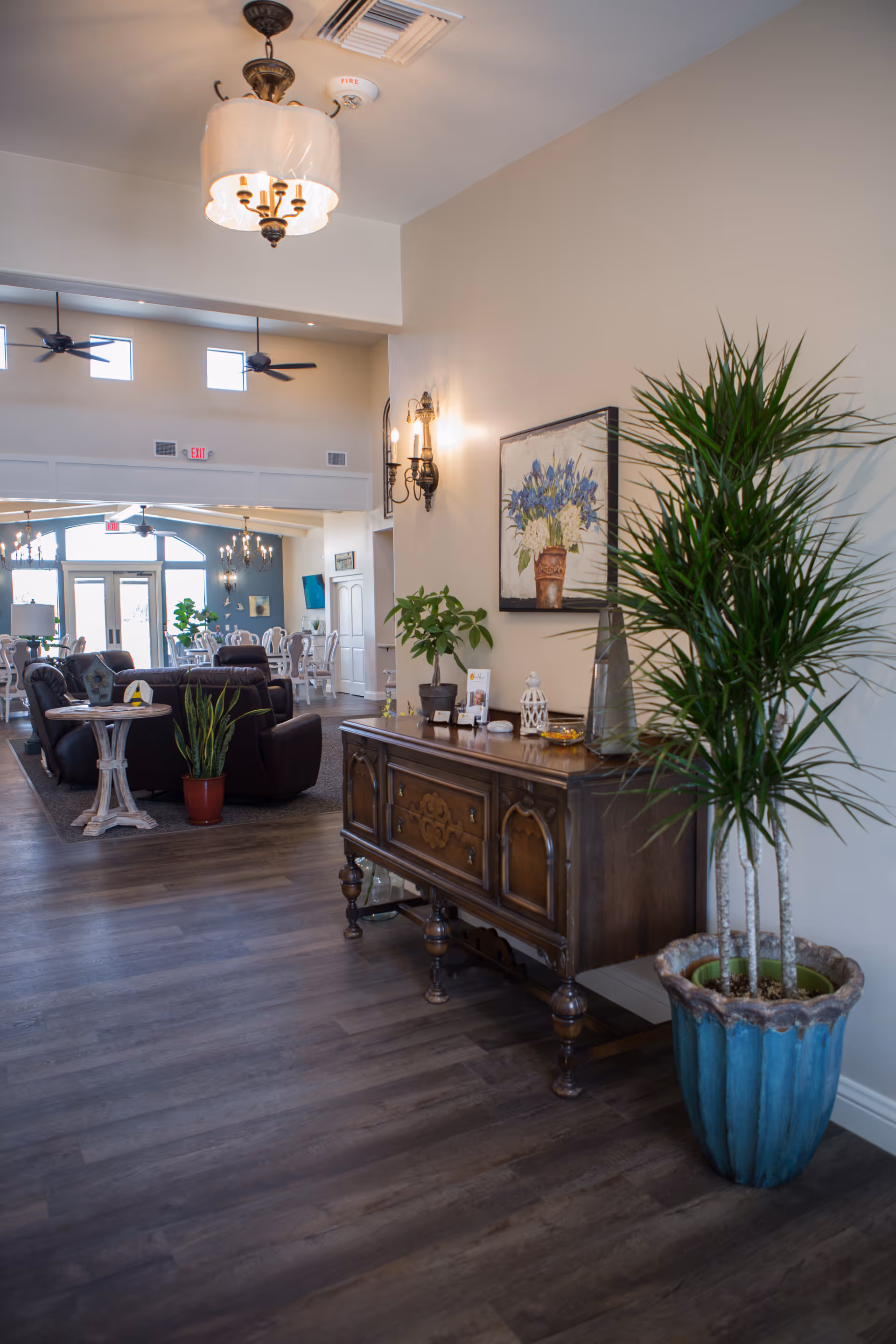 Interior view of a senior living facility showing a hallway with wooden flooring, a vintage wooden sideboard with decorative items and plants on top, a large potted plant in a blue pot, and a painting of flowers on the wall. In the background, there is a seating area with dark armchairs, tables, and ceiling fans, leading to a dining area with white chairs and tables near large windows.