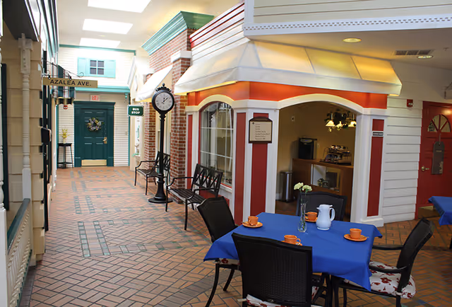Indoor hallway designed to look like a small town street with storefronts, benches, a clock, and street signs including Azalea Ave and Bus Stop. A table with a blue tablecloth, four chairs, orange cups, and a white pitcher is in the foreground.