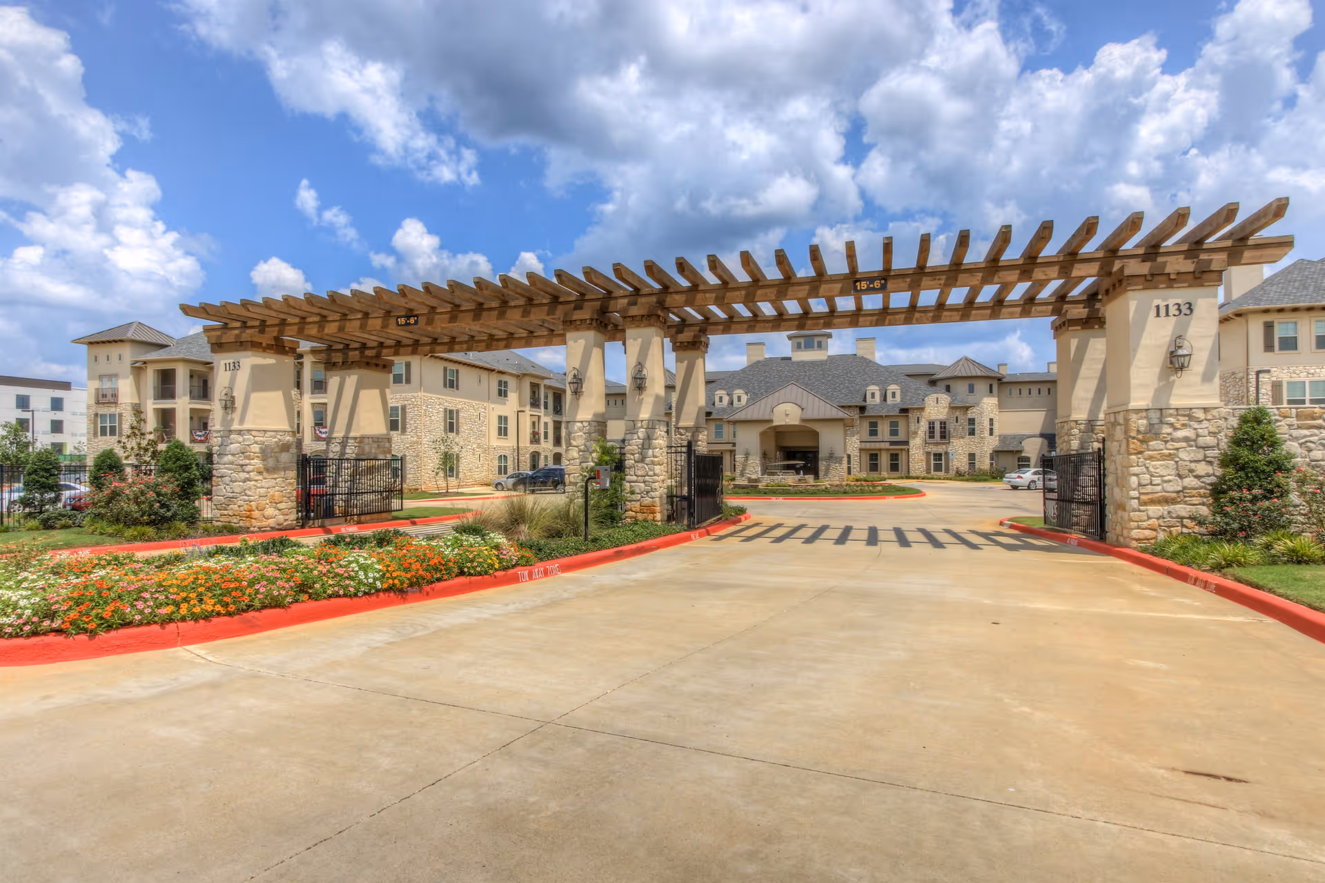 Entrance to a senior living facility with a large wooden pergola structure supported by stone pillars. The driveway is lined with red curbs and colorful flower beds. The building in the background is multi-story with beige walls, stone accents, and multiple windows under a partly cloudy sky.