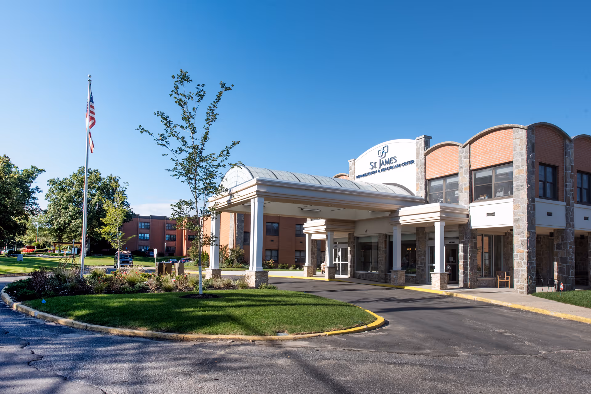 Exterior view of St. James Rehabilitation & Healthcare Center building with a covered entrance, a flagpole with an American flag, trees, and a clear blue sky.
