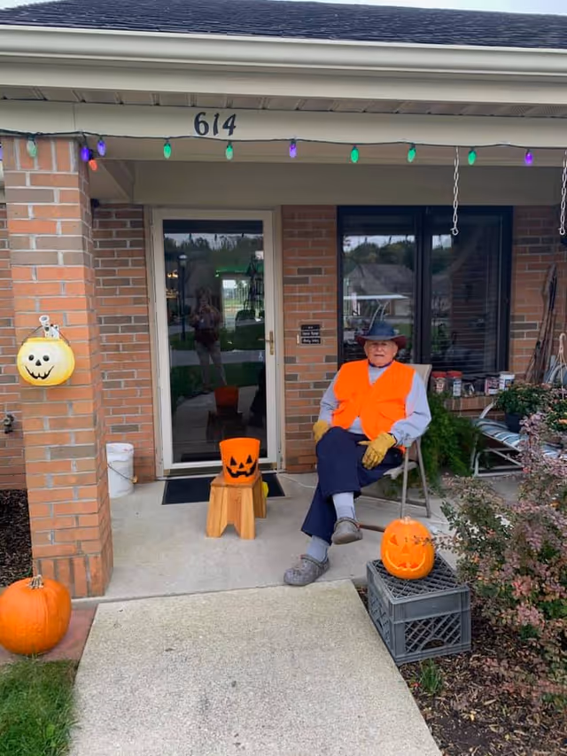 An elderly man wearing an orange safety vest, gray shirt, blue pants, gloves, and a black hat is sitting on a chair on the front porch of a brick building. The porch is decorated with Halloween items including carved pumpkins, a pumpkin bucket, and a hanging pumpkin decoration. The building number 614 is visible above the door, and colorful string lights hang from the porch roof.