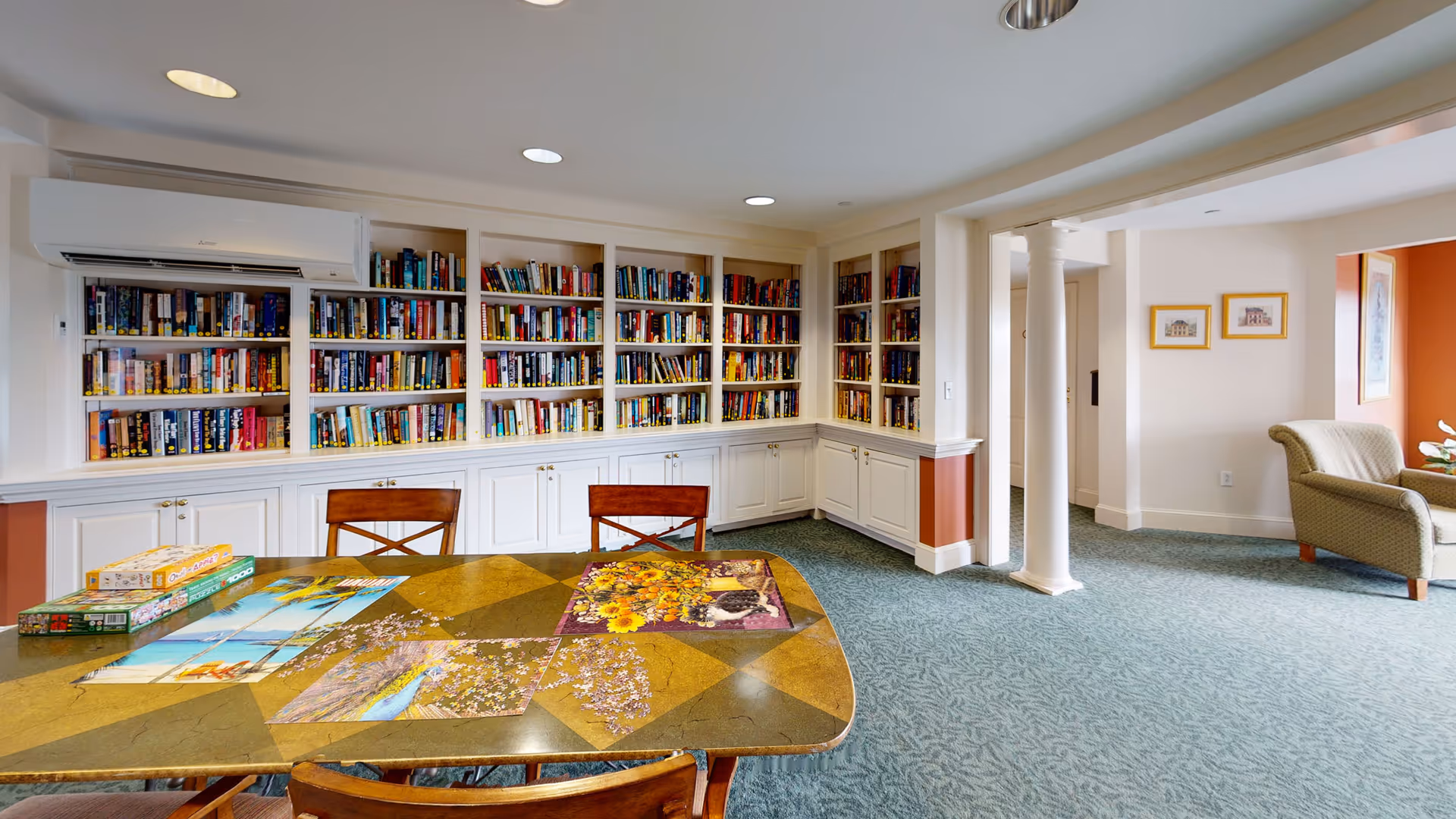 A cozy reading and activity room with a large built-in bookshelf filled with books along the back wall. In front of the bookshelf is a table with several puzzles and chairs around it. The room has carpeted flooring, white walls with some orange accents, and a comfortable armchair near a window letting in natural light.