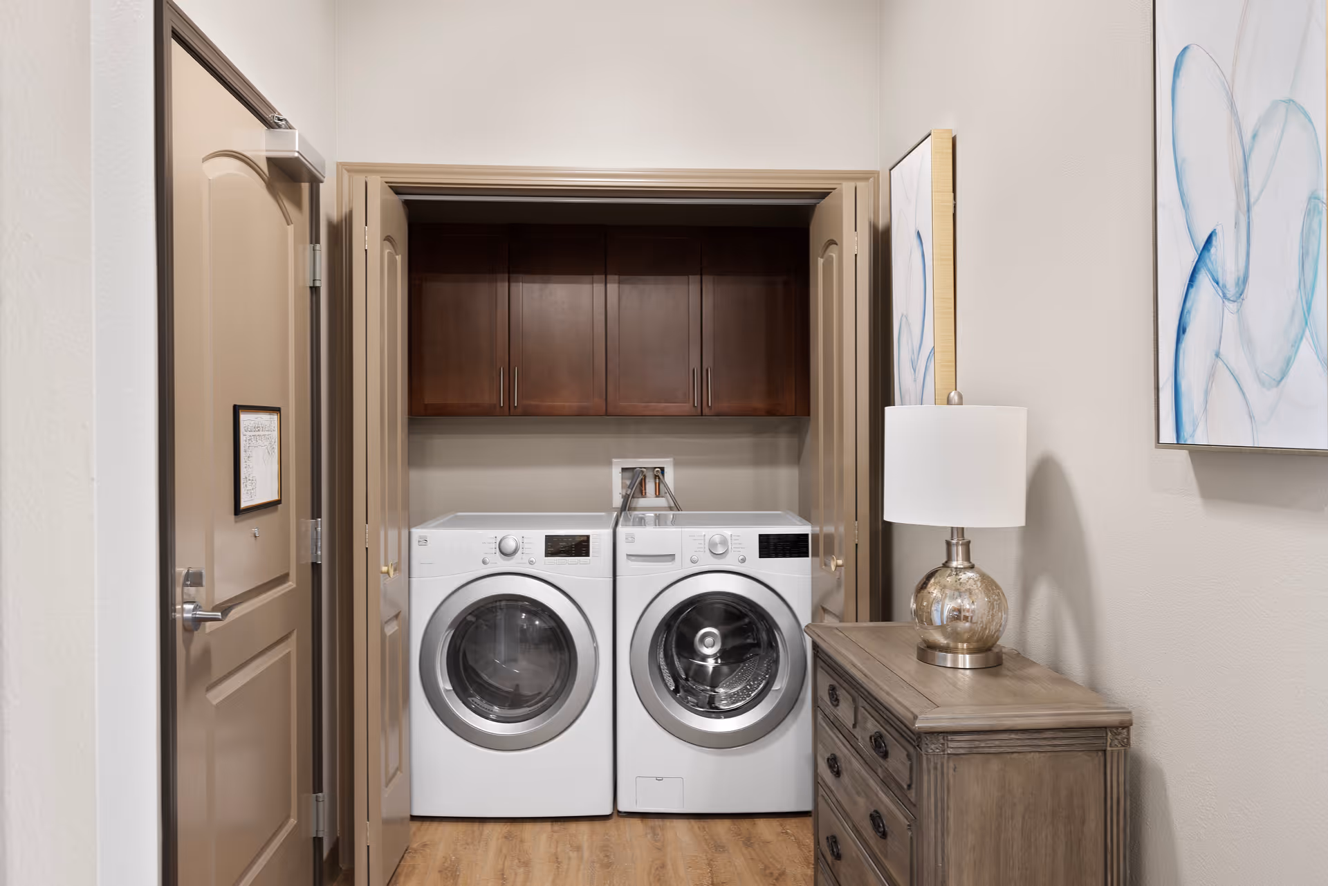 A laundry area with a front-loading washing machine and dryer side by side inside a closet with double doors. Above the machines are dark wooden cabinets. To the right of the closet is a wooden dresser with a glass table lamp and a framed abstract artwork on the wall.