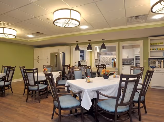 Dining room with several tables covered in white tablecloths, wooden chairs with blue cushions, pendant lights, and a serving kitchen area.