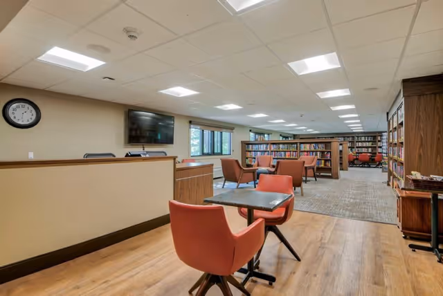 Interior view of a senior living facility library or common area with wooden flooring, several bookshelves filled with books, multiple seating areas with orange chairs and small tables, a wall-mounted TV, and a clock on the wall.