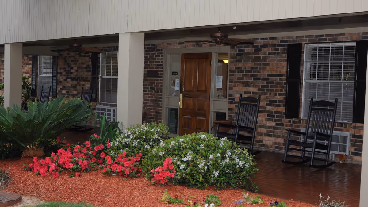 Front porch area of a brick building with a wooden door, two windows with black shutters, and a covered patio featuring black rocking chairs. There are green shrubs with white and red flowers and a small palm plant in front of the porch.
