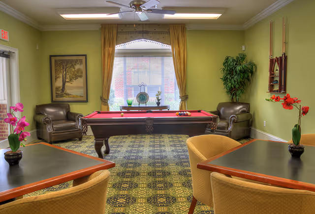 Communal game room with a red-felt pool table centered under a ceiling fan, surrounded by armchairs, tables with flowers, and a window with drapes.